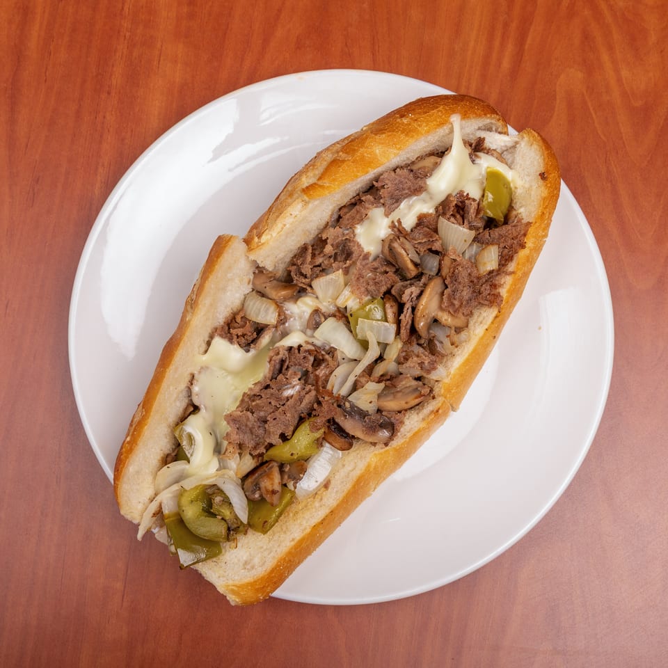 Cheesesteak sandwich with sliced beef, melted cheese, mushrooms, onions, and green peppers on a white plate.