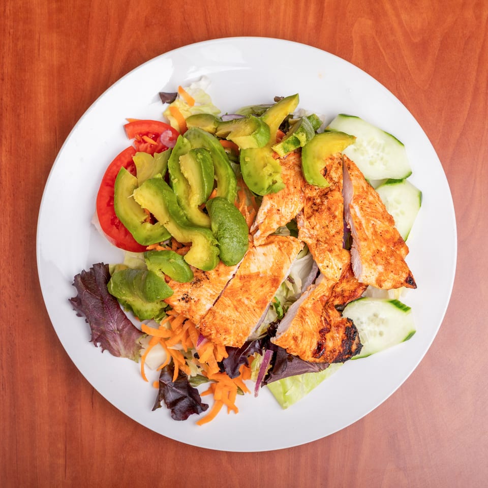 Grilled chicken salad topped with avocado, tomato, cucumber, carrots, and mixed greens on a white plate.