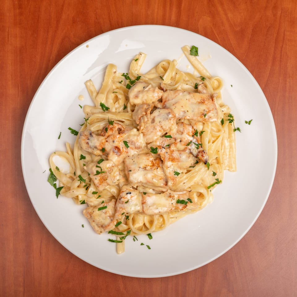Creamy fettuccine Alfredo topped with grilled chicken and parsley, served on a white plate atop a wooden table.