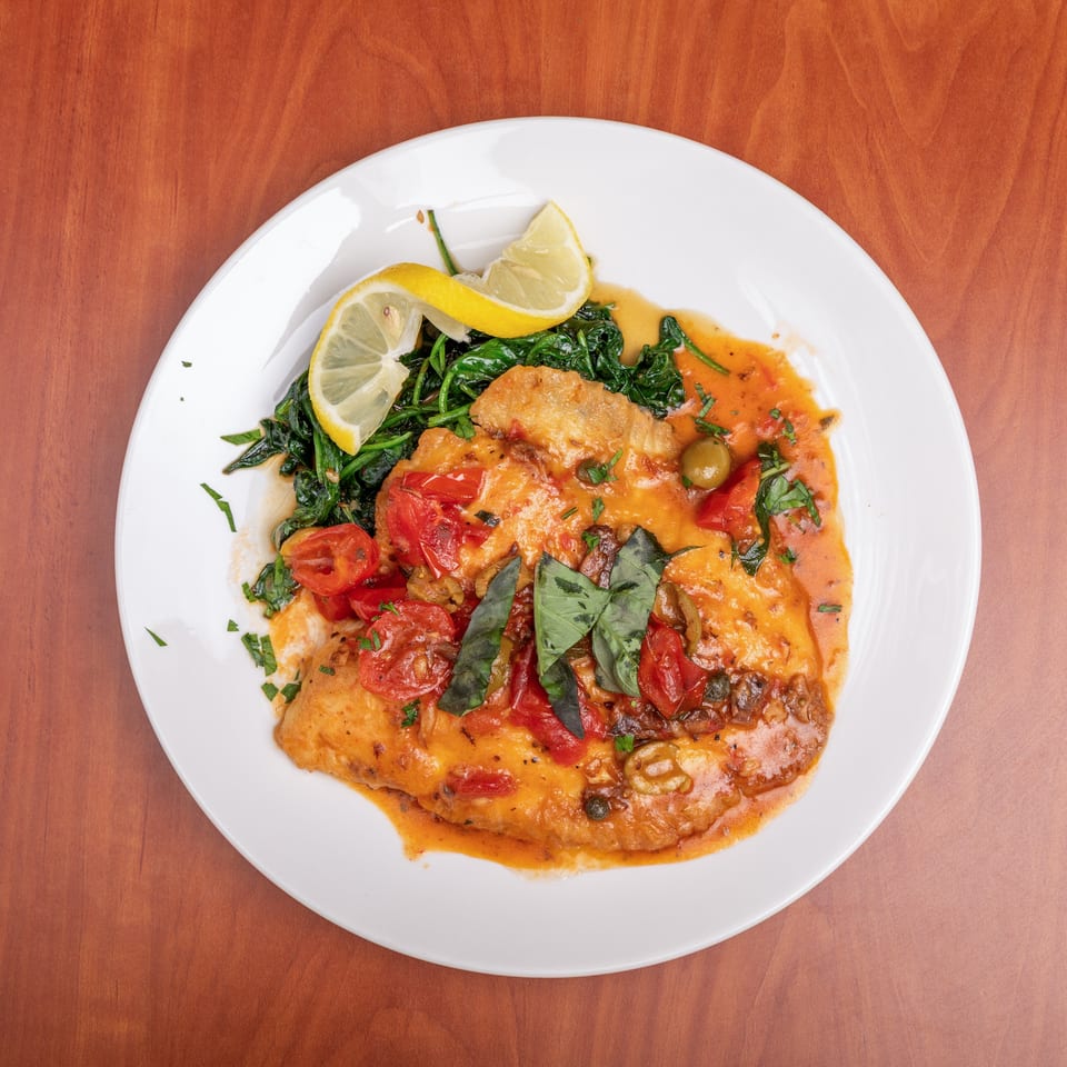 Breaded fish fillets in tomato sauce with olives, tomatoes, basil, sautéed spinach, and lemon wedges on a white plate.