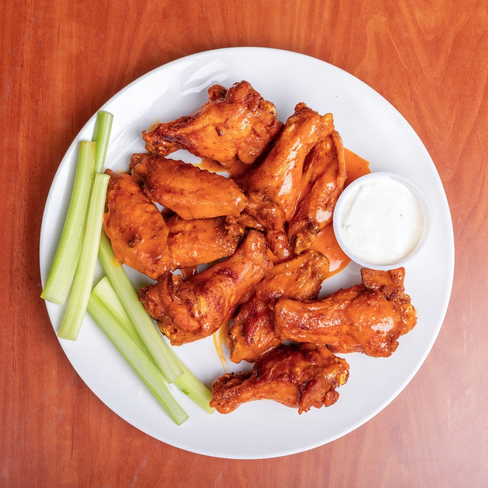 Buffalo chicken wings with celery sticks and ranch dressing on a white plate atop a wooden table.