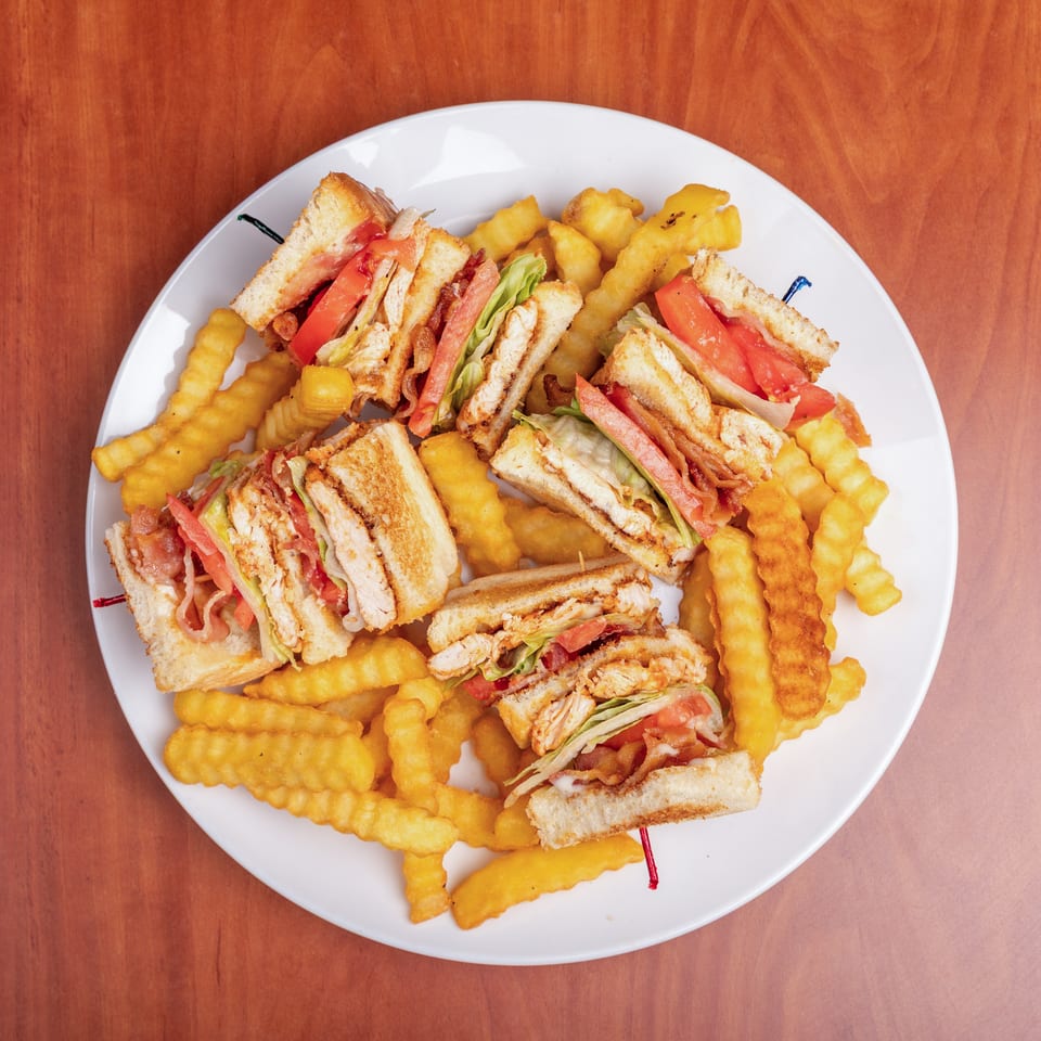 Plate of club sandwiches with chicken, bacon, lettuce, and tomato surrounded by crinkle-cut fries on a wooden table.