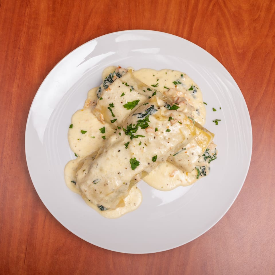White plate of ravioli covered in creamy sauce, spinach, and chopped parsley on a wooden table.