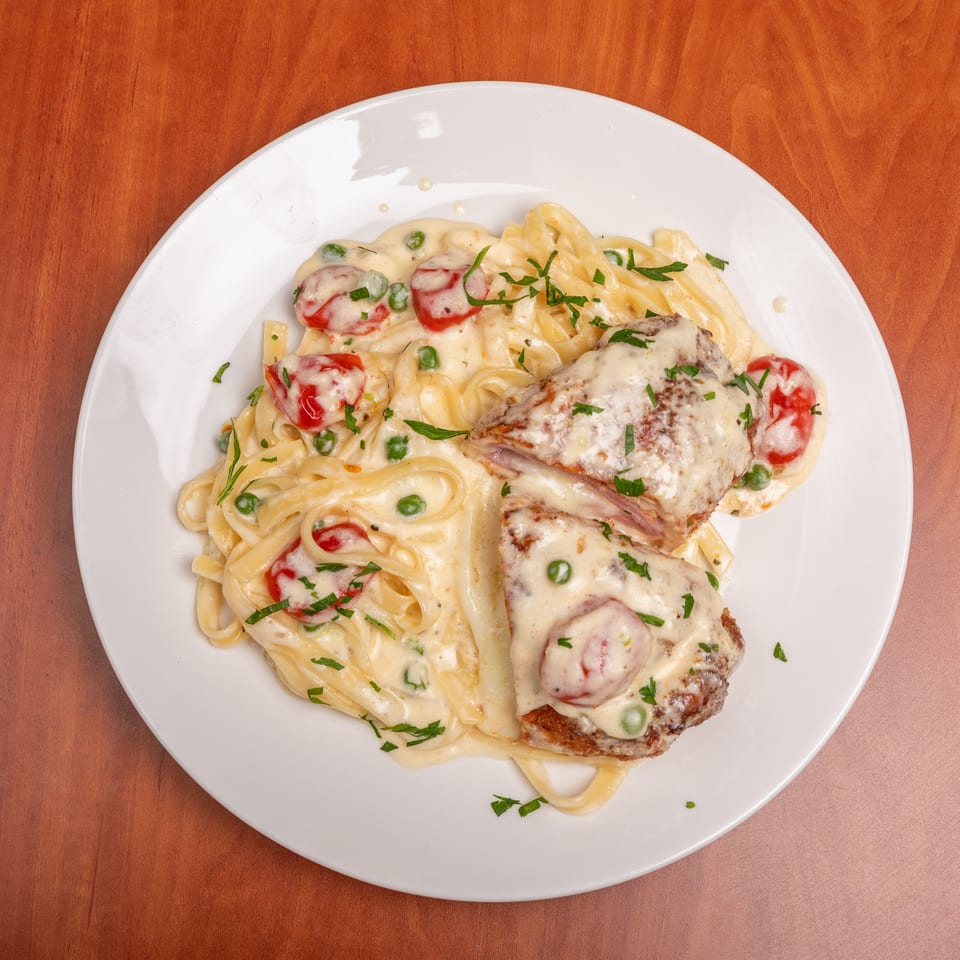 Creamy fettuccine with peas and tomatoes, topped with sliced grilled chicken, melted cheese, and parsley on a white plate.